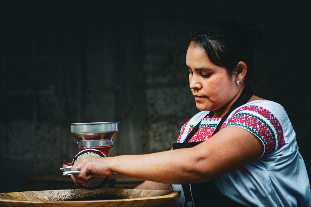A woman in traditional attire grinds coffee beans using a manual grinder in a dark setting.